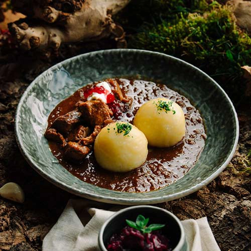 Das leckere Gulasch wird auf einem Teller serviert zusammen mit zwei Knödeln. Der Teller steht auf einem Waldboden.