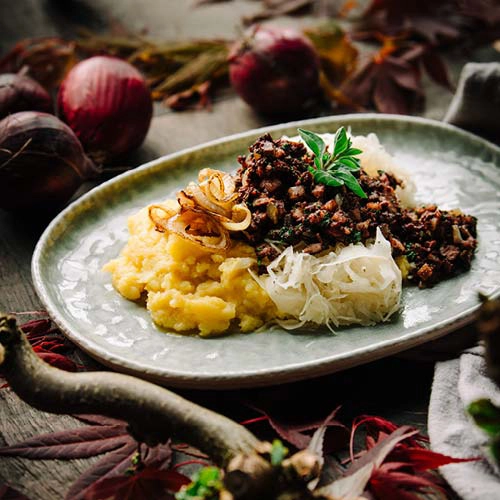 Ein rustikales Gericht mit Rotwurst, Sauerkraut, Kartoffelbrei und knusprigen Röstzwiebeln – angerichtet auf einem ovalen Teller. Drumherum Rohstoffe auf einem Holzboden.