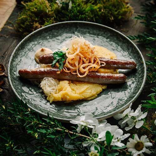 2 Thüringer Bratwürsten, Sauerkraut, Kartoffelbrei und Senf sind angerichtet auf einen dunkelgrünen Teller. Im Hintergrund ist ein Waldboden, Blüten und Moos.