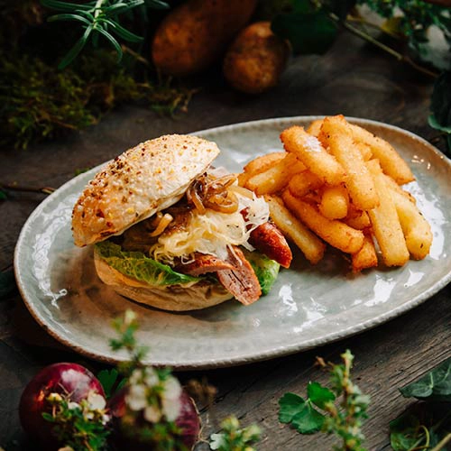 Ein vegetarischer Burger mit gebranntem Ziegenkäse, Preiselbeeren, frischem Grünzeug, Thymian und Honig. Dazu Kloßpommes auf einem Teller angerichtet.