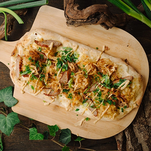 Eine leckere Pinsa mit Thüringer Rostbratwurst, Sauerkraut und einem Hauch Senf. Angerichtet auf deinem Holzbrett. Daneben Dekorationen wie Efeu und Baumrinde.