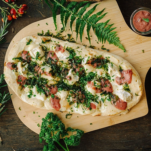 Die Rennsteig Pinsa „Ziegenglück“ ist mit Ziegenkäse, Preiselbeeren und frittiertem Rucola. Angerichtet auf einem Holzbrett. Drumherum Walddekrationen, wie Beeren & Moos.