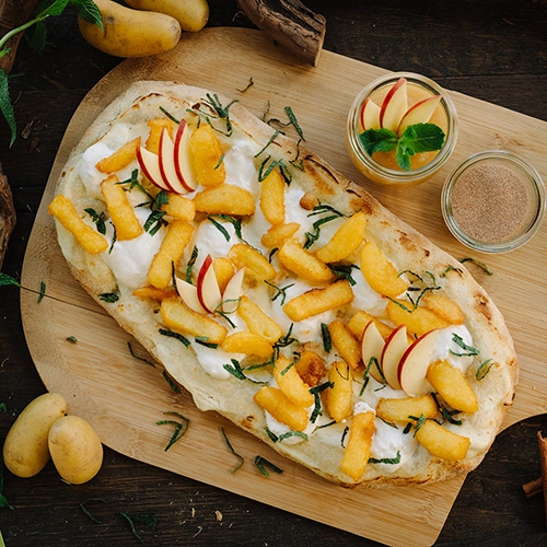 Eine süße Pinsa mit gebratenen Kloßpommes, Schmand, Minze und frischen Äpfeln – angerichtet auf einem Holzbrett. Drumherum Schalen mit Apfelmuss, Zucker&Zucker.