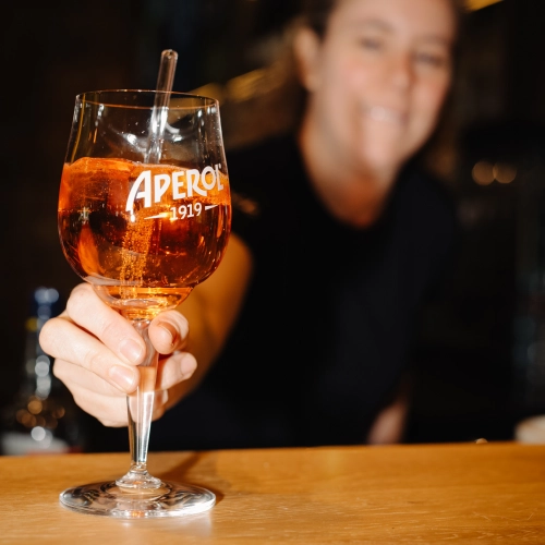 Eine Frau serviert ein Glas Aperol. Die Frau ist im Hintergrund unscharf. Der Fokus auf dem Bild ist auf dem Aperol.