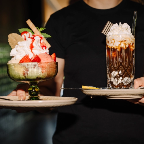 Frau trägt ein Eiskaffe und ein Erdbeerbecher mit Sahne zu einem Tisch.