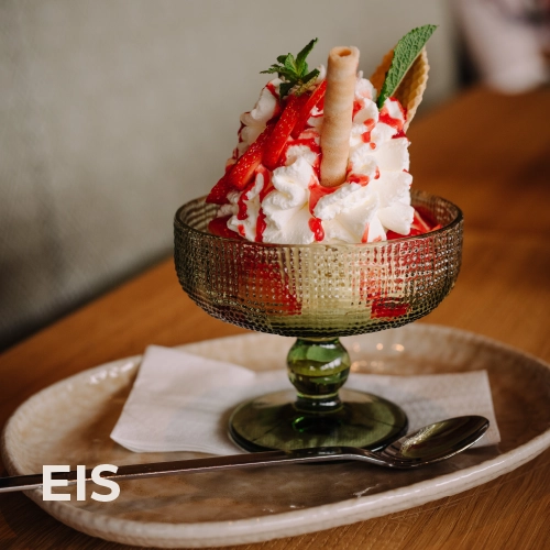 Das Titelbild für die Menürubrik „Kuchen & Co.“: Das ansprechend angerichtete Erdbeereis. Sahne und frische Erdbeeren sowie eine Waffelrolle schmücken den Eisbecher.