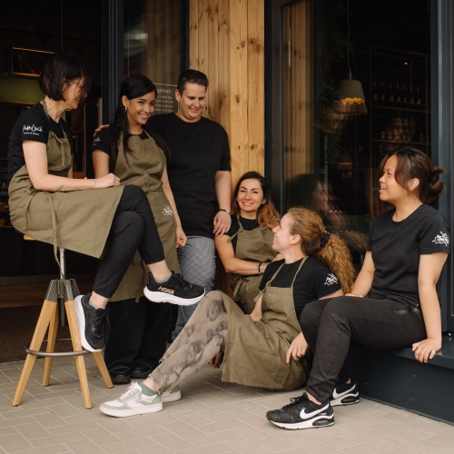 Das Team sitzt vor dem Restaurant zusammen. Eine Person sitzt auf dem Boden, die anderen stehen und eine sitzt auf dem Barhocker.